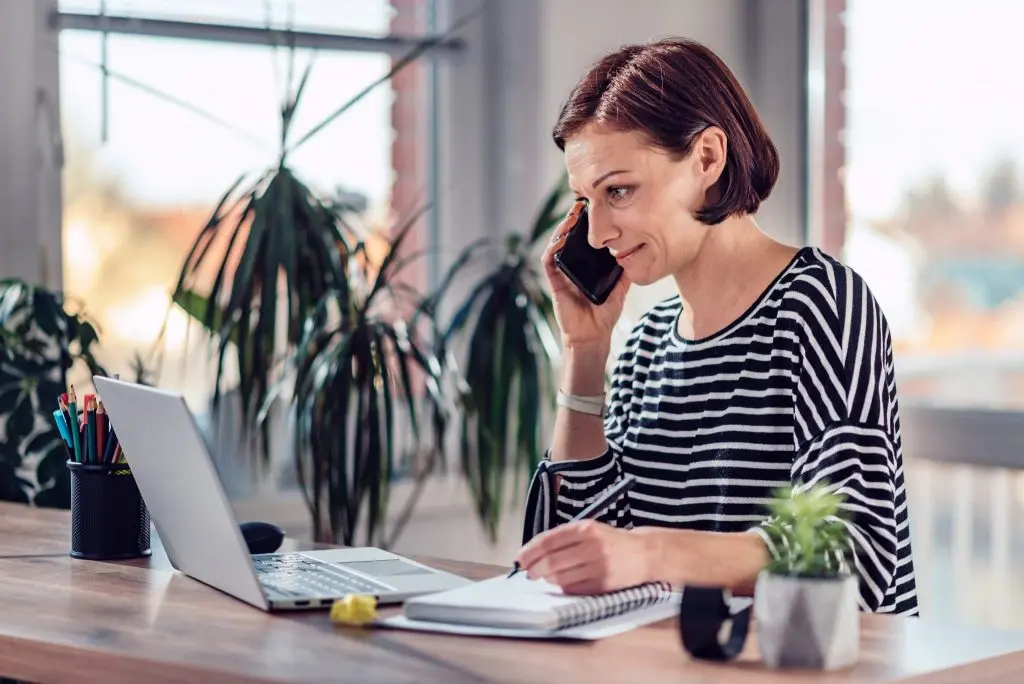 Woman talking on the smart phone and writing notes in notebook in the office
