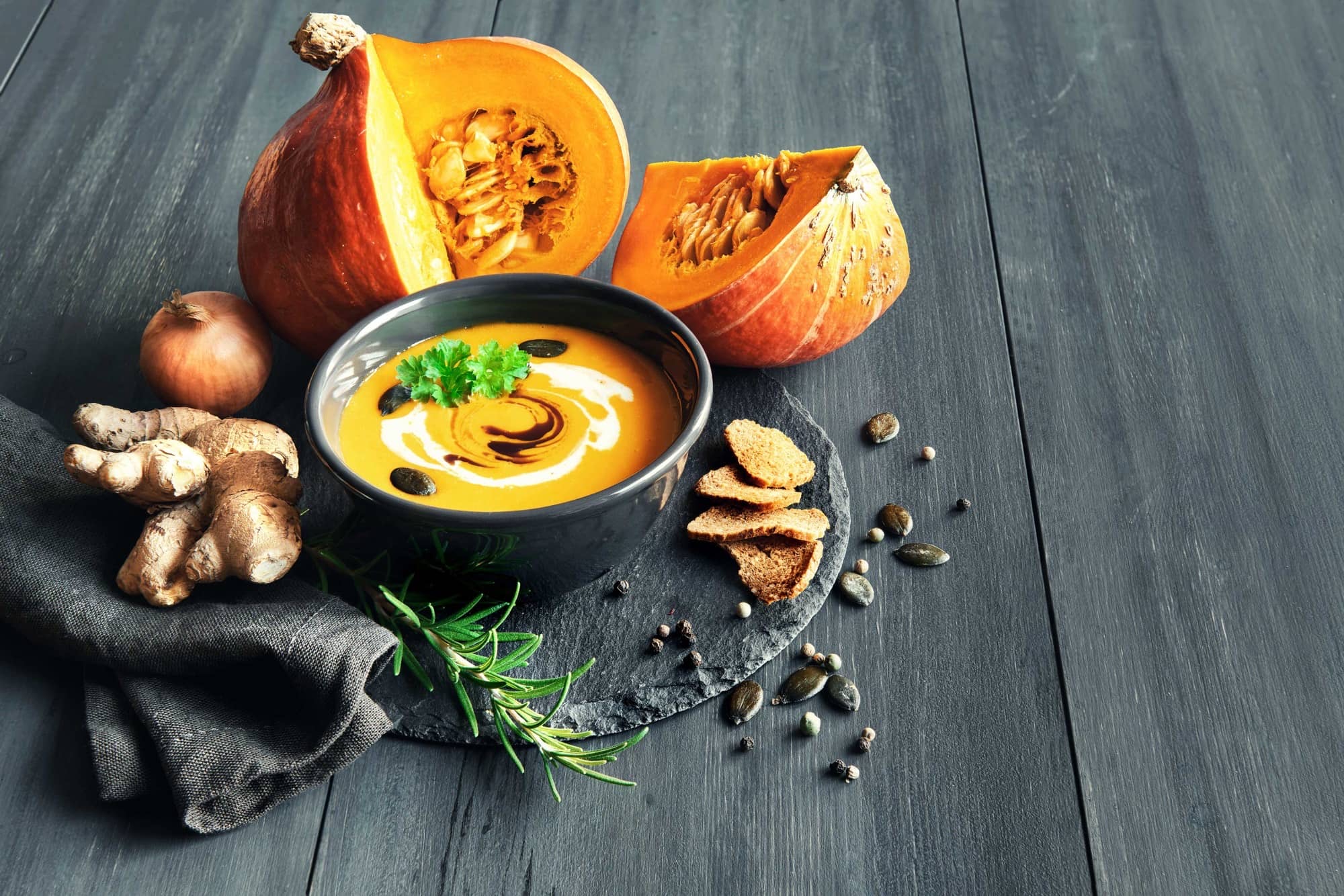 Crema de carbassa amb gingebre i crostons d’herbes en un bol negre, acompanyada de carbassa tallada, cebolla, gingebre i crostons d’herbes sobre una taula de fusta