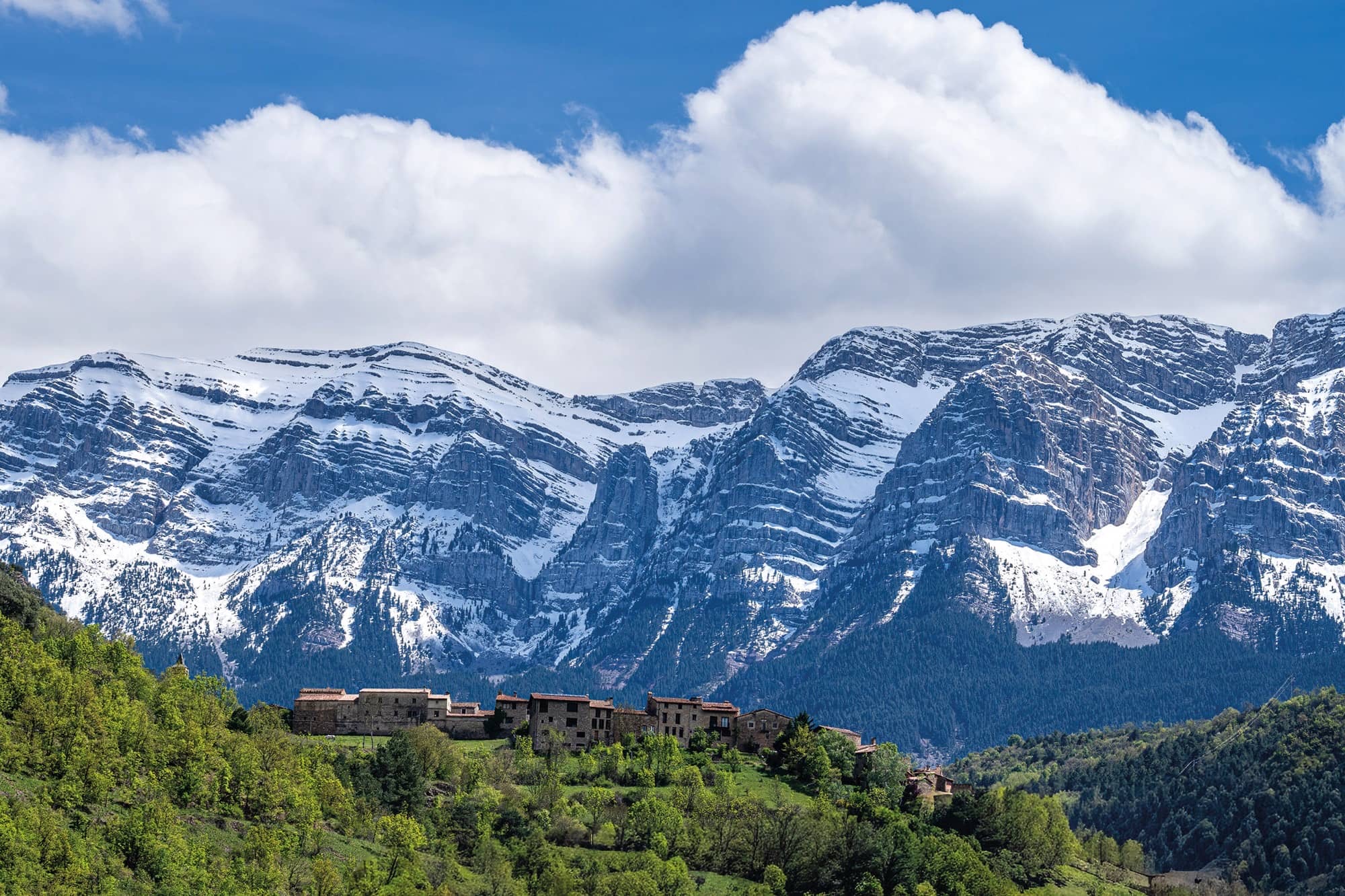 Paisatge de muntanya amb boscos, un nucli de poblament tradicional i les cimeres rocoses i nevades del Cadí al fons.