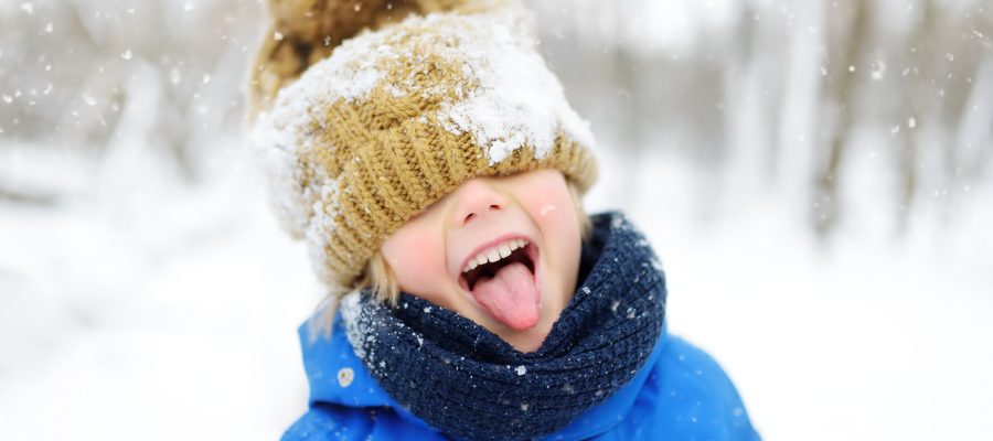 Funny little boy in blue winter clothes walks during a snowfall. Outdoors winter activities for kids. Cute child wearing a warm hat low over his eyes catching snowflakes with his tongue
