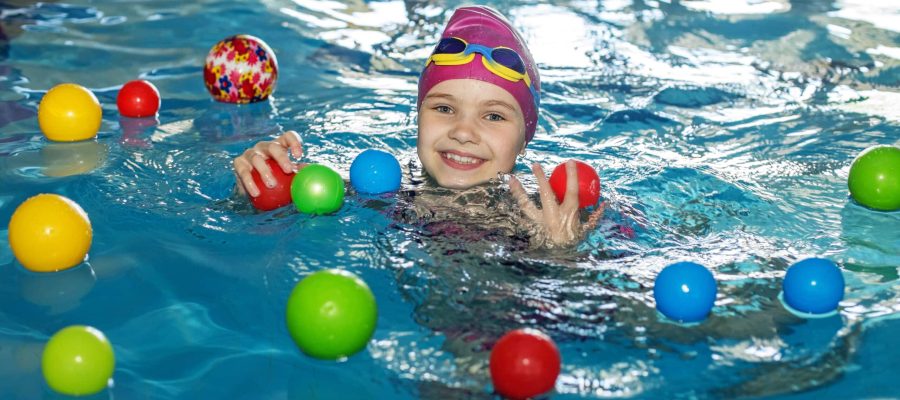 Happy child schoolgirl is studying at swimming lessons in pool. Swimming cap and goggles. Lots of colorful balls. Concept of healthy lifestyle and sports.